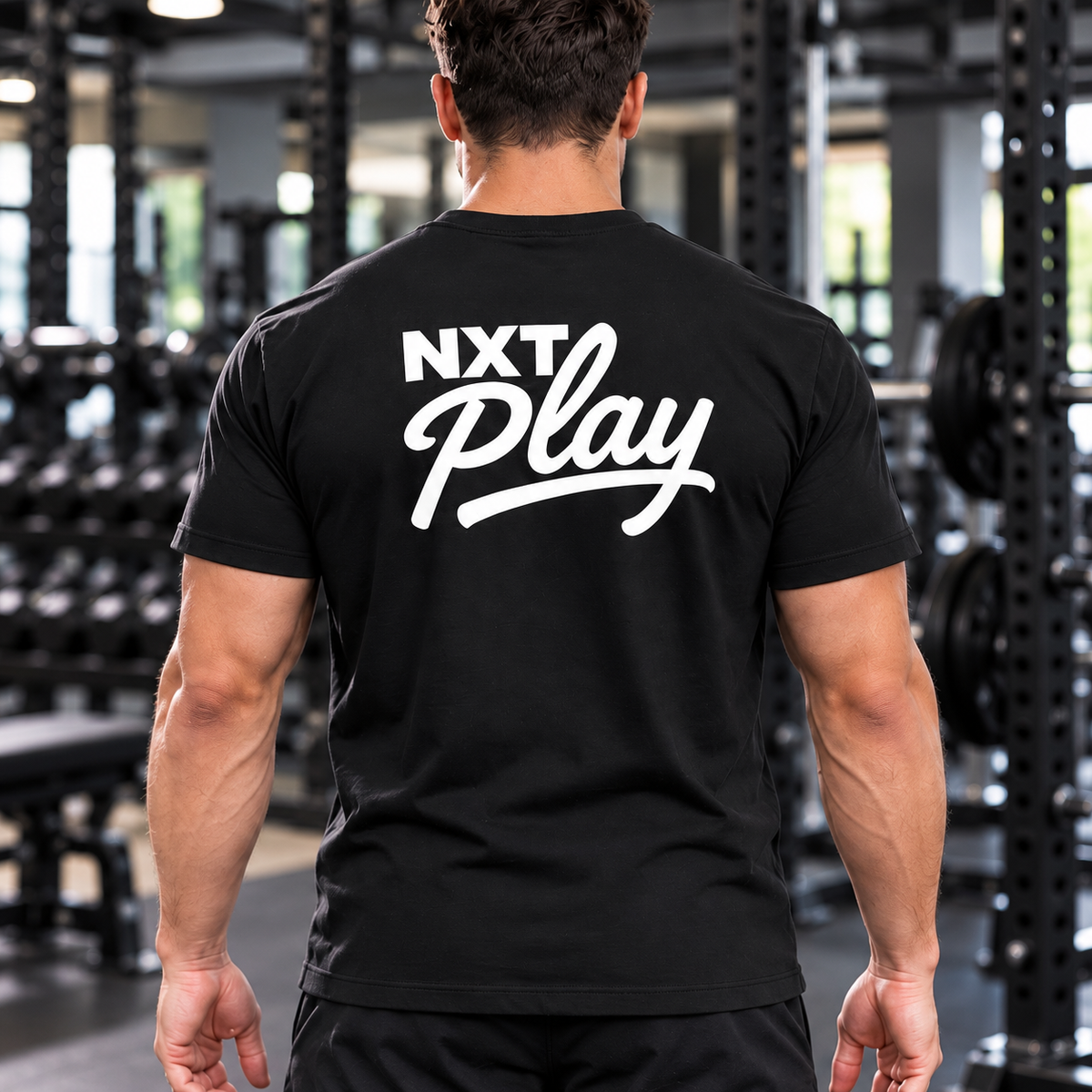 A man viewed from behind in a gym, wearing a black t-shirt with a large white 'NXT Play' logo printed across the back.