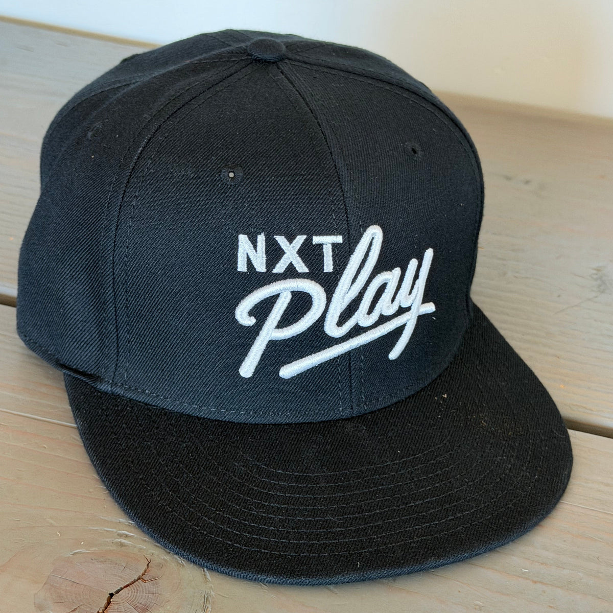 A black snapback cap with white embroidered text reading 'NXT Play' on the front