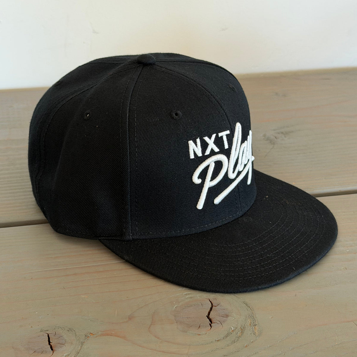 A black flat-brim snapback cap with white embroidered text reading 'NXT Play' on the front