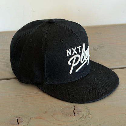 A black flat-brim snapback cap with white embroidered text reading 'NXT Play' on the front