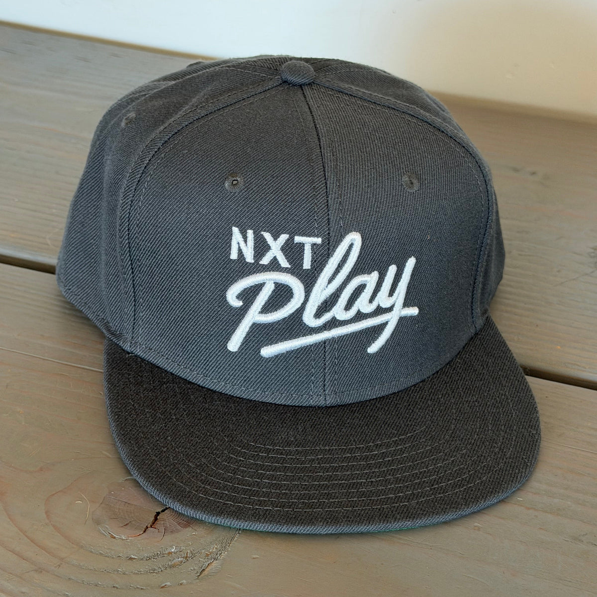 A dark gray snapback cap with 'NXT Play' embroidered in white on the front