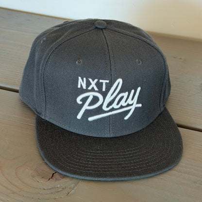 A dark gray snapback cap with 'NXT Play' embroidered in white on the front