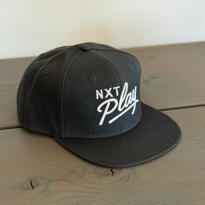 A black snapback cap with 'NXT Play' embroidered in white on the front