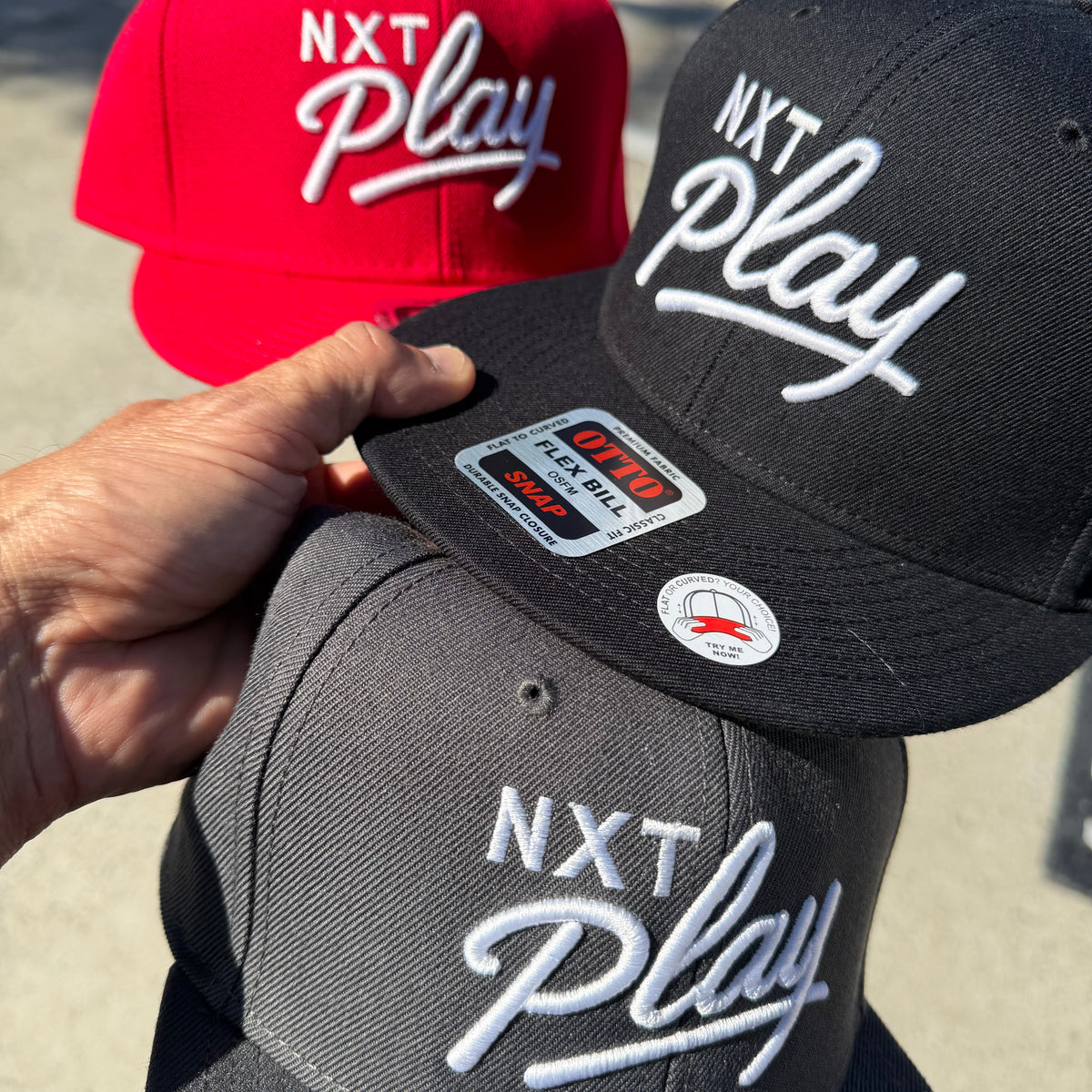 Three snapback caps with 'NXT Play' embroidered on the front are stacked outdoors. A hand is holding the top black cap, with a red cap in the background and a dark gray cap in the foreground.