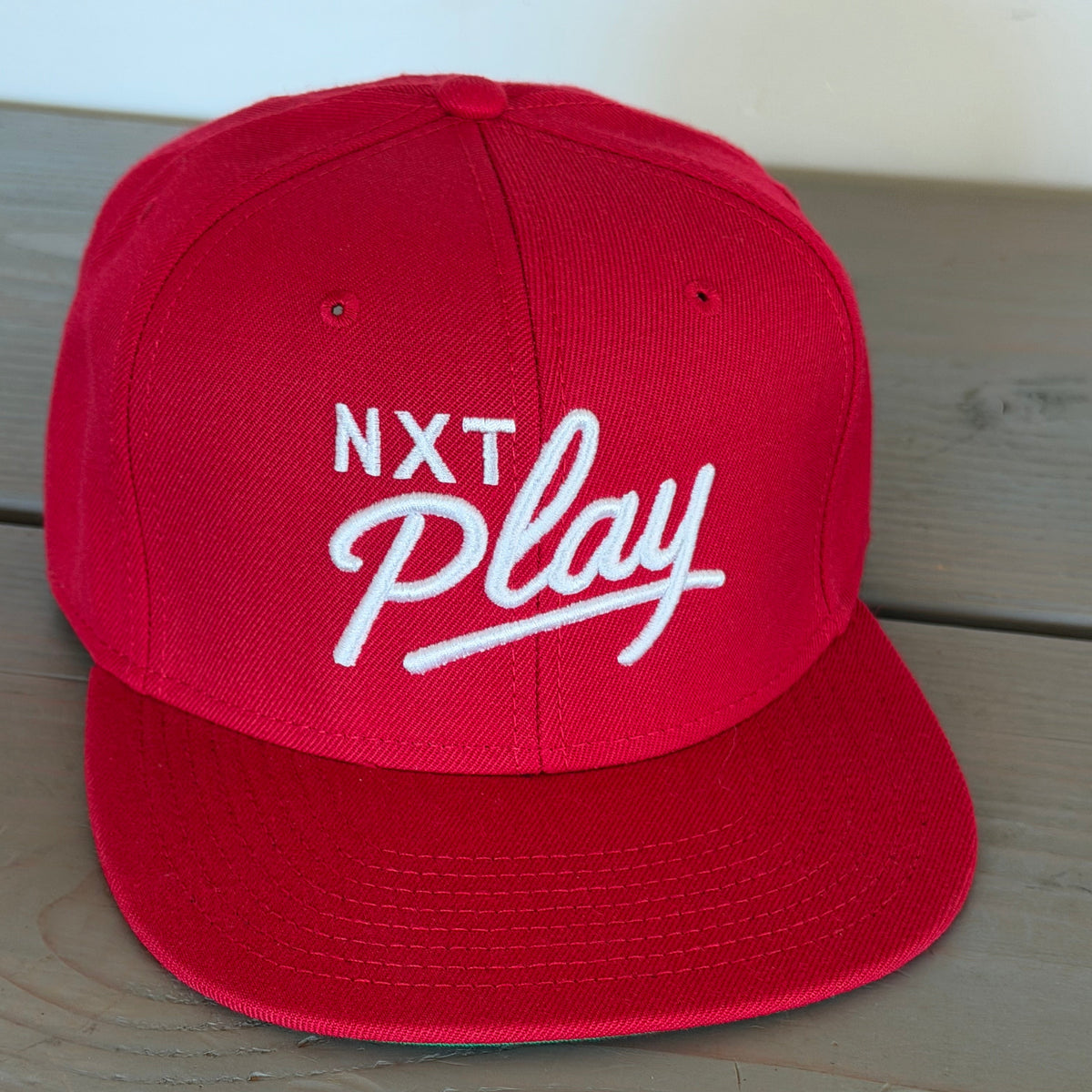 A red flat-brim baseball cap with 'NXT Play' embroidered in white on the front