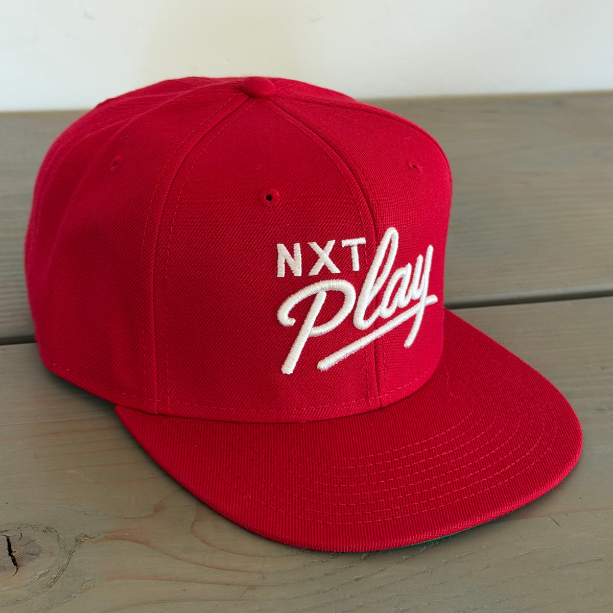 A red flat-brim baseball cap with white embroidered text reading 'NXT Play' on the front
