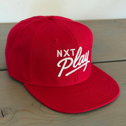 A red flat-brim baseball cap with white embroidered text reading 'NXT Play' on the front